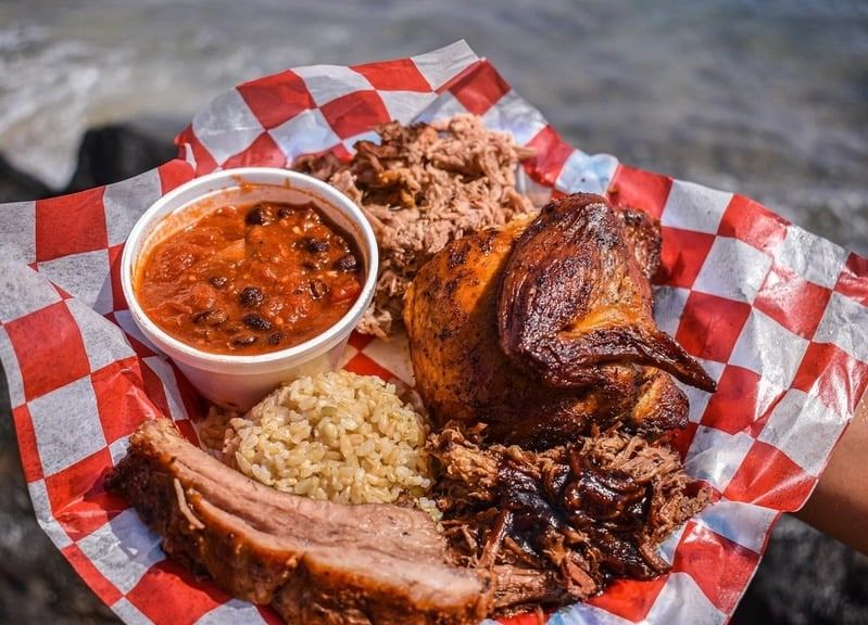 A plate of smoked chicken, ribs and brisket with beans and rice on red checkered paper