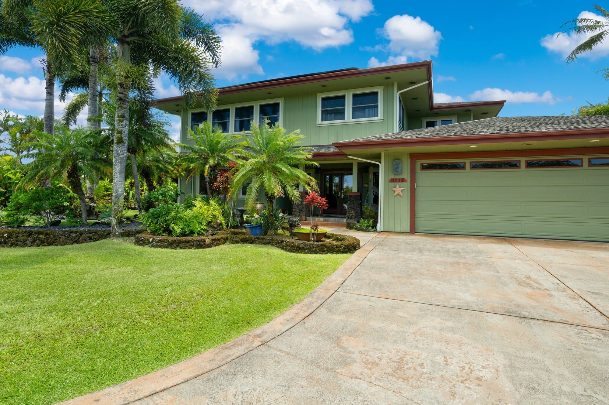 The two-story green house with a garage, a wide lawn and tall palms
