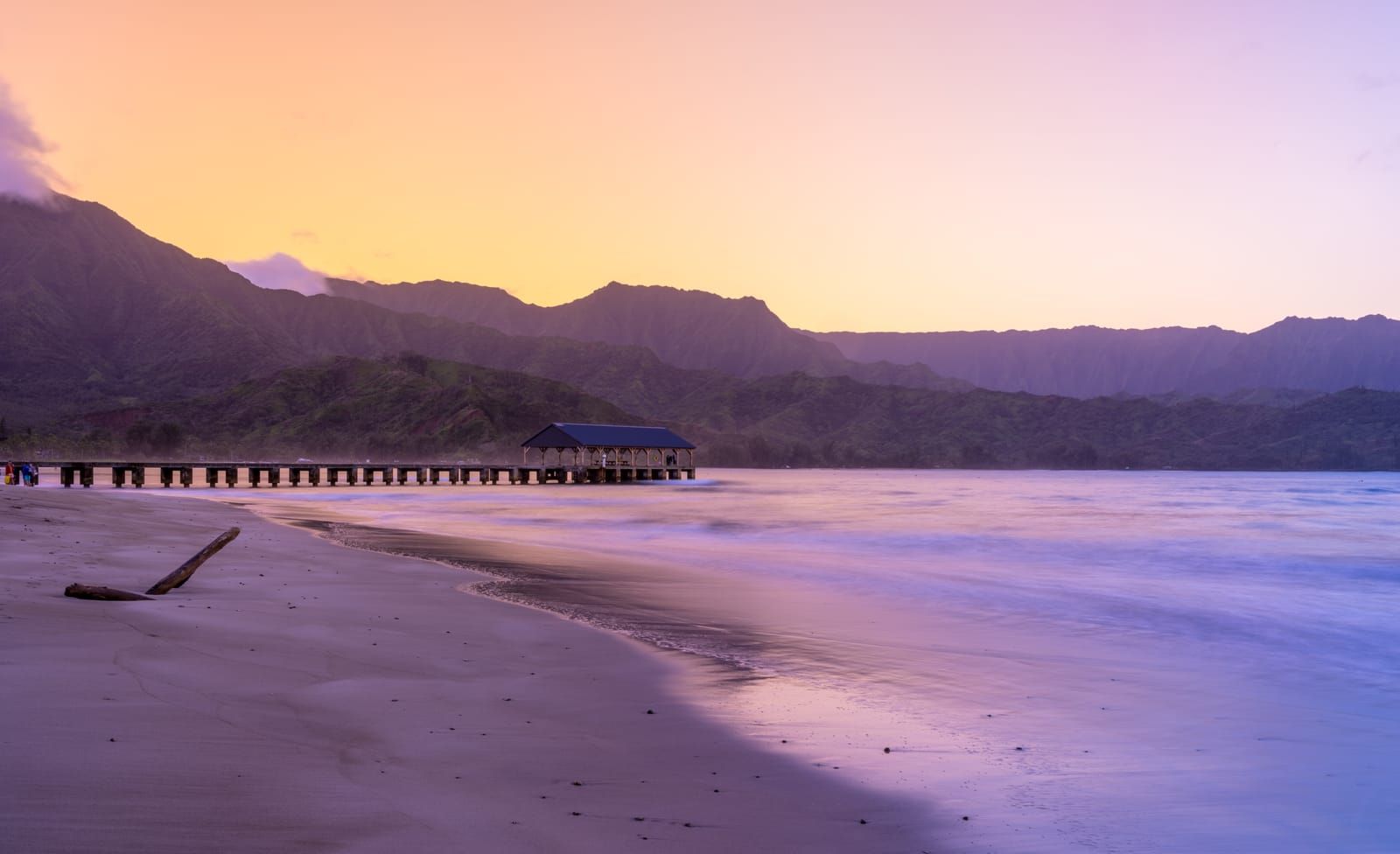 Hanalei Pier at dawn under a pink and gold sky, with mountains behind