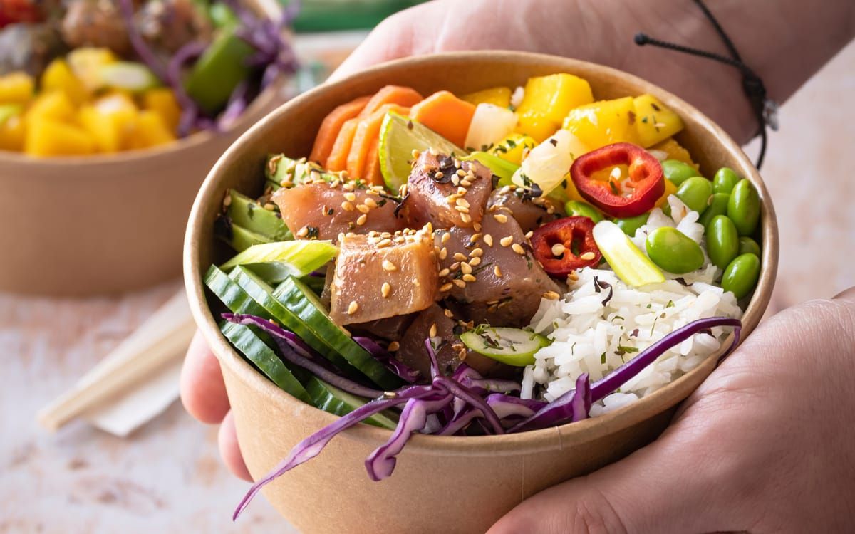 Hands holding a takeaway bowl of ahi poke with rice and vegetables