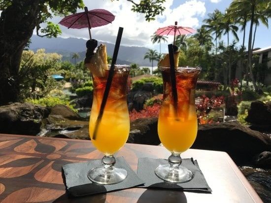 Two tropical cocktails with paper umbrellas on a table overlooking palms and mountains